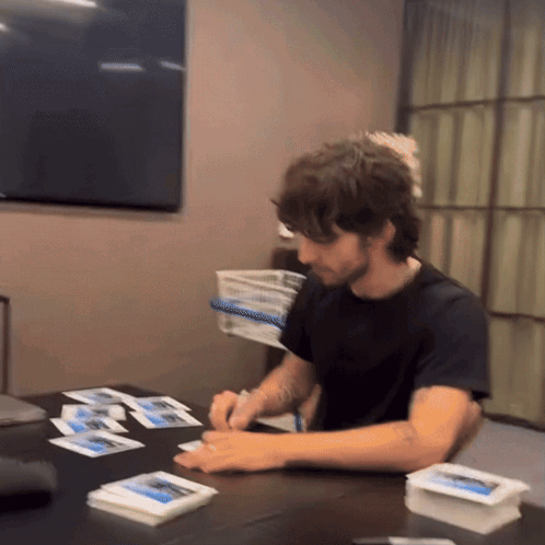 A Man In A Black Shirt Sits At A Table With A Bunch Of Cards On It GIF