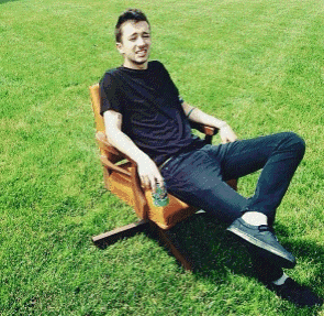 A Man In A Black Shirt Sits In A Wooden Chair In The Grass GIF