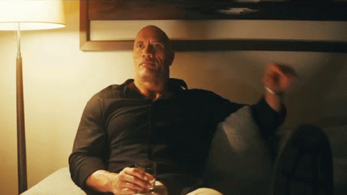 A Man In A Black Shirt Sits On A Couch Holding A Glass Of Whiskey GIF