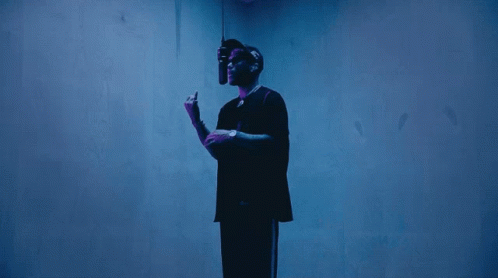 A Man In A Black Shirt Stands In Front Of A Blue Wall Holding A Microphone GIF