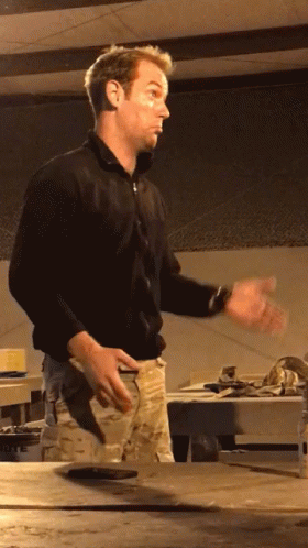 A Man In A Black Shirt Stands In Front Of A Table With His Hands Outstretched GIF