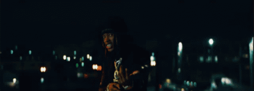 A Man In A Black Shirt With A Skull On It Is Standing In The Dark GIF