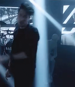 A Man In A Black Shirt With The Word Bas On The Back Is Standing In A Dark Room . GIF