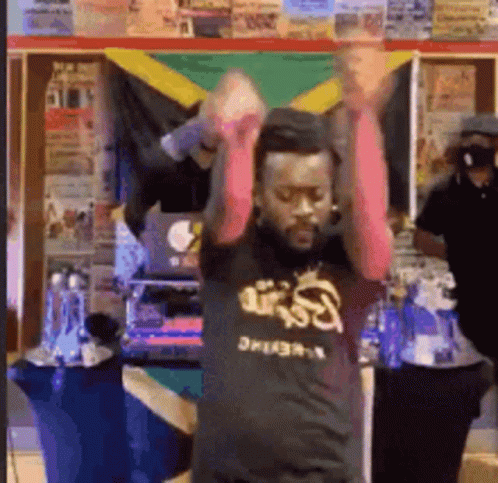 A Man In A Black Shirt With The Word Reggae On It Is Dancing In Front Of A Jamaican Flag . GIF