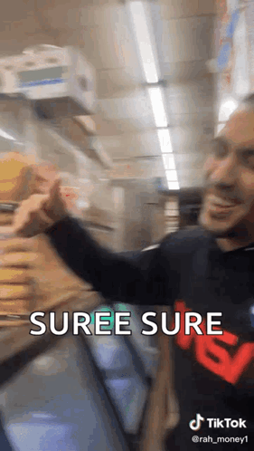 A Man In A Black Shirt With The Word Suree Sure On It Is Standing In Front Of A Refrigerator In A Store . GIF