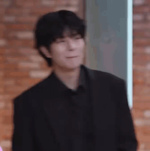 A Man In A Black Suit And Black Shirt Is Making A Funny Face While Standing In Front Of A Brick Wall . GIF