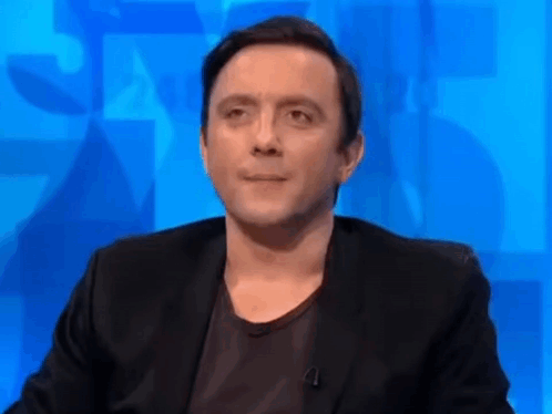A Man In A Black Suit And Black Shirt Is Sitting In Front Of A Blue Wall . GIF