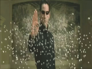 A Man In A Black Suit And Sunglasses Is Standing In Front Of A Bunch Of Bullets . GIF