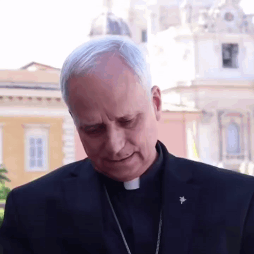 A Man In A Black Suit Has A Badge On His Lapel That Says ' Cardinal ' On It GIF