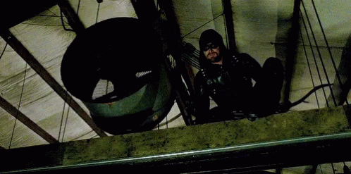 A Man In A Black Suit Is Holding A Bow And Arrow While Sitting On A Ledge . GIF