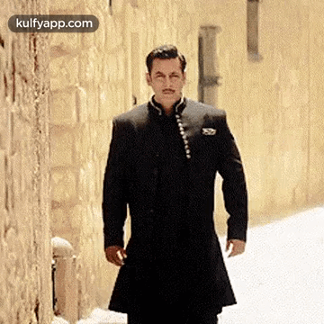A Man In A Black Suit Is Standing In Front Of A Brick Wall . GIF