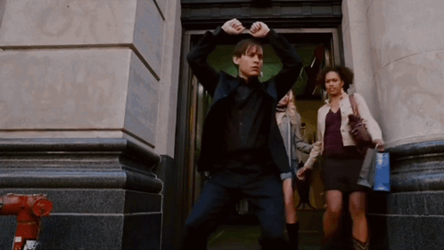 A Man In A Black Suit Is Standing In Front Of A Building GIF