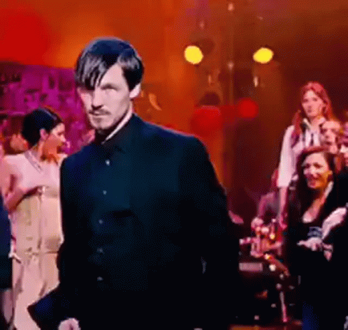 A Man In A Black Suit Is Standing In Front Of A Group Of People . GIF