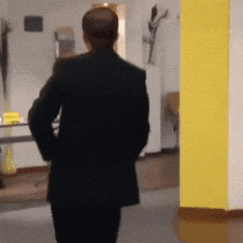 A Man In A Black Suit Is Walking In A Hallway With His Hands In His Pockets GIF