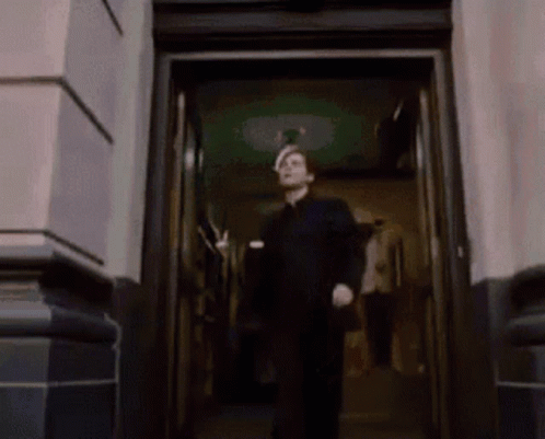 A Man In A Black Suit Is Walking Out Of A Doorway GIF