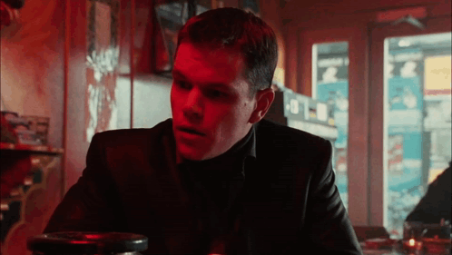 A Man In A Black Suit Sits At A Table In A Restaurant GIF