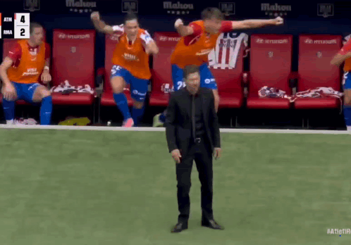 A Man In A Black Suit Stands In Front Of A Group Of Soccer Players On The Field GIF