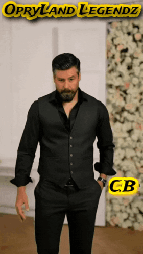 A Man In A Black Suit Stands In Front Of A Wall Of Flowers With Opryland Legendz Written On The Bottom GIF
