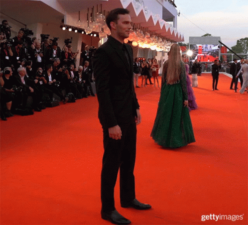 A Man In A Black Suit Stands On A Red Carpet With A Woman In A Green Dress Standing Behind Him GIF