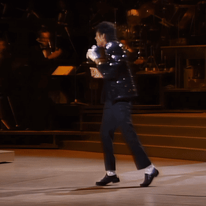 A Man In A Black Suit Stands On A Stage Holding A Microphone GIF
