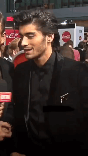 A Man In A Black Suit Talks Into A Microphone In Front Of A Coca-Cola Sign GIF
