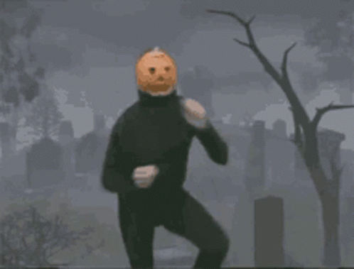 a man in a black suit with a pumpkin on his head is standing in a cemetery .