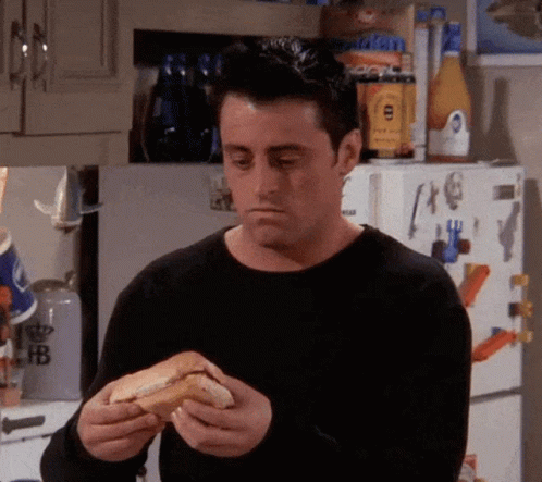 A Man In A Black Sweater Is Eating A Hot Dog In Front Of A Refrigerator In A Kitchen . GIF