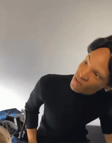 A Man In A Black Sweater Is Sitting On A Bed And Looking At Something GIF