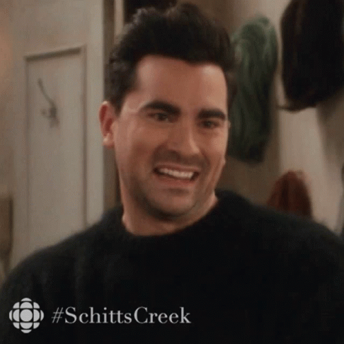 A Man In A Black Sweater Is Smiling In A Room With A Sign That Says Schitts Creek . GIF