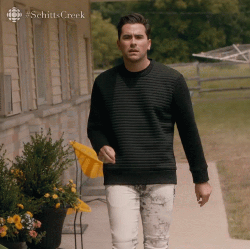 A Man In A Black Sweater Is Walking Down A Sidewalk In Front Of A House With #Schittscreek On The Bottom GIF