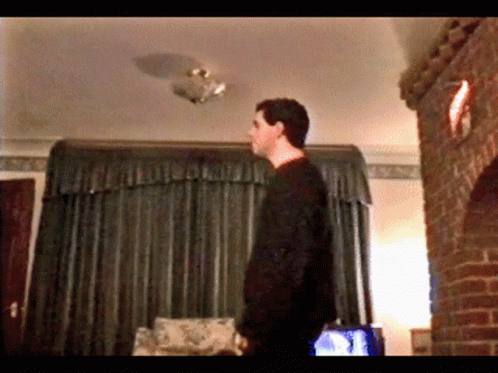 A Man In A Black Sweater Stands In A Living Room Looking Up At The Ceiling GIF
