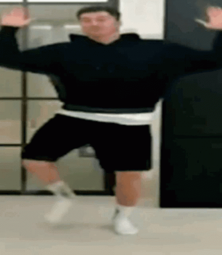 A Man In A Black Sweatshirt And Black Shorts Is Dancing In A Room . GIF
