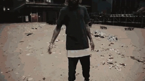 A Man In A Black T-Shirt Is Standing In A Dirt Area GIF