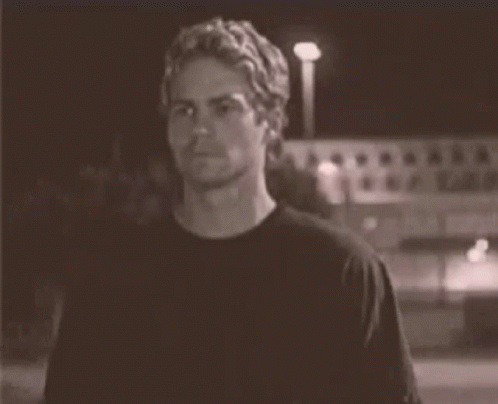 A Man In A Black T-Shirt Is Standing In Front Of A Building At Night . GIF