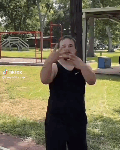 A Man In A Black Tank Top And Black Shorts Is Standing In A Park With His Hands On His Face . GIF