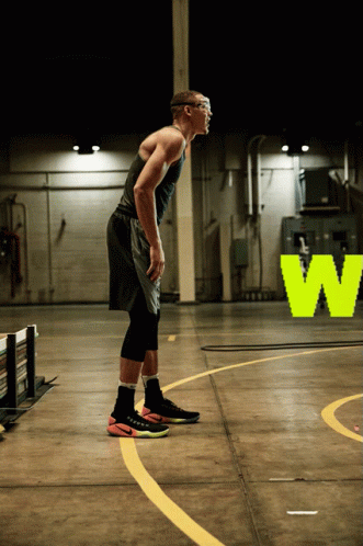 A Man In A Black Tank Top And Black Shorts Stands On A Basketball Court In Front Of A Neon W Sign GIF