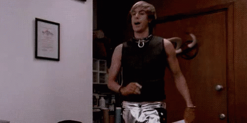 A Man In A Black Tank Top And Silver Shorts Is Standing In Front Of A Door . GIF