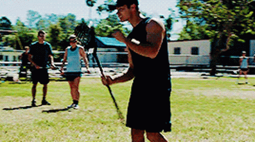 A Man In A Black Tank Top Is Holding A Lacrosse Stick In A Field GIF