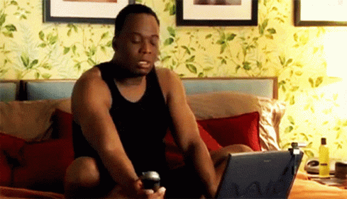 A Man In A Black Tank Top Is Sitting On A Bed Using A Laptop GIF