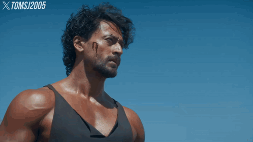 A Man In A Black Tank Top Stands In Front Of A Blue Sky With Toms 2005 Written On The Bottom Right GIF