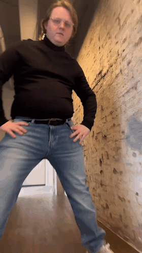 A Man In A Black Turtleneck And Blue Jeans Stands With His Hands In His Pockets GIF