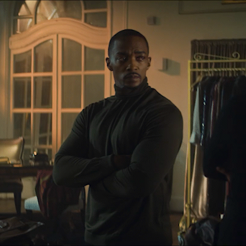 A Man In A Black Turtleneck Stands With His Arms Crossed In A Dark Room GIF