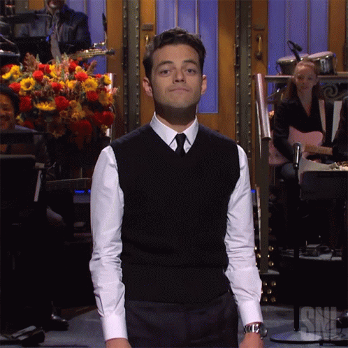 A Man In A Black Vest And White Shirt Stands In Front Of A Snl Sign GIF