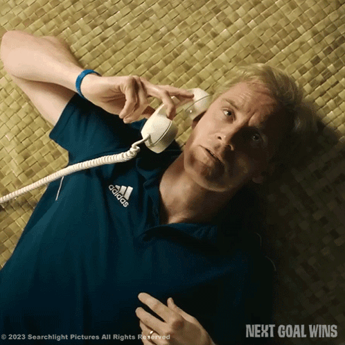 A Man In A Blue Adidas Shirt Is Laying On The Floor Talking On A Telephone GIF