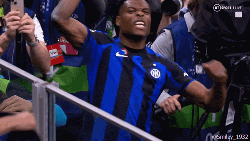 A Man In A Blue And Black Striped Shirt Stands In Front Of A Bt Sport 1 Live Camera GIF