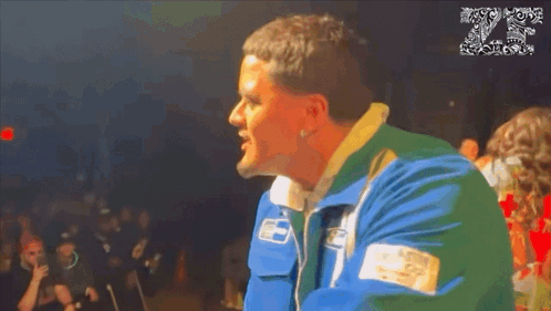 A Man In A Blue And Green Jacket Stands In Front Of A Crowd With The Number 20 On The Bottom GIF