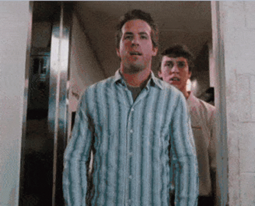 A Man In A Blue And White Striped Shirt Stands In A Doorway GIF