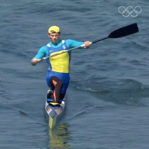 A Man In A Blue And Yellow Outfit Is Paddling A Canoe In The Water With The Olympic Rings In The Background GIF