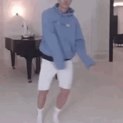 A Man In A Blue Hoodie And White Shorts Is Standing In Front Of A Piano In A Living Room . GIF