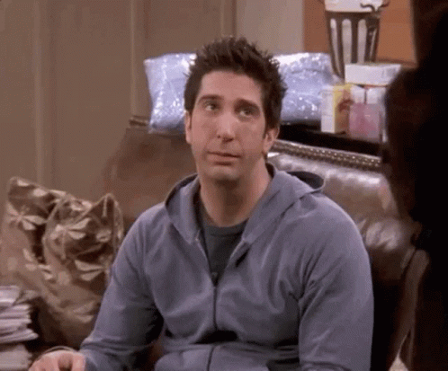 A Man In A Blue Hoodie Is Sitting On A Couch And Looking At The Camera . GIF